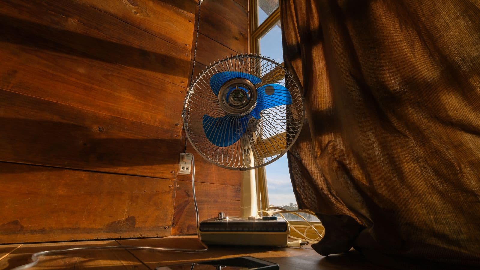 A vintage electric fan with blue blades beside a curtained window in a wood-paneled room in warm evening light.