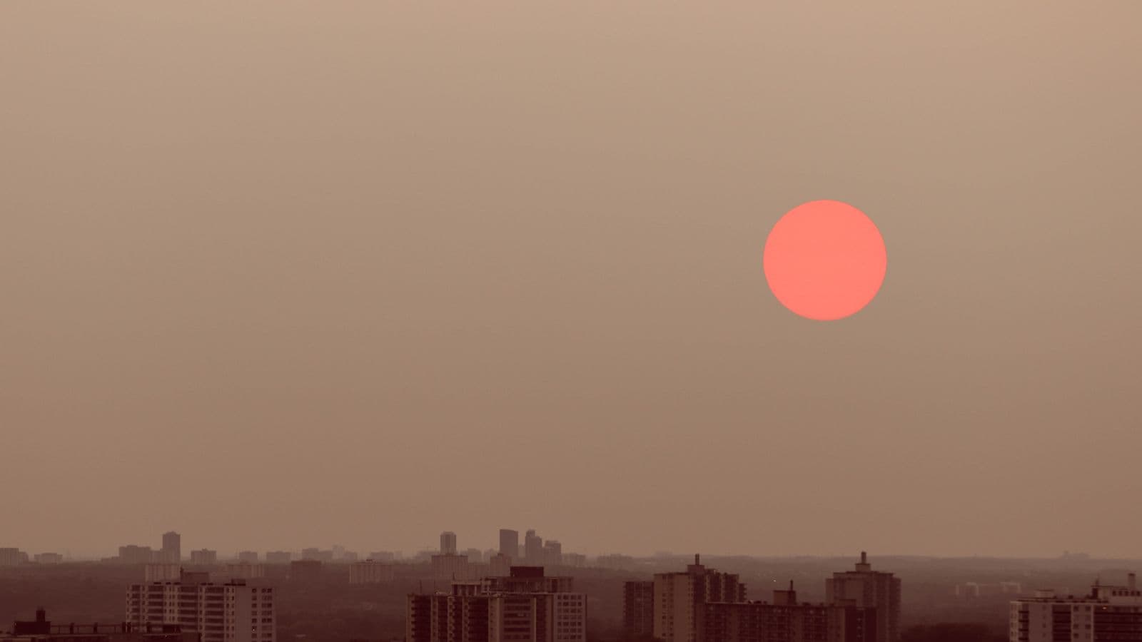 A deep-red sun glowing through thick brown smoky haze above a silhouetted city skyline.
